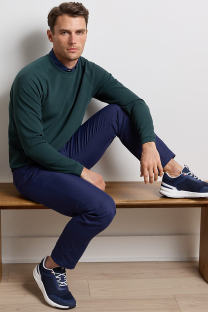A man sits on a wooden bench wearing a dark green mock-neck long-sleeve shirt, navy blue trousers, and navy blue athletic sneakers against a light gray wall.