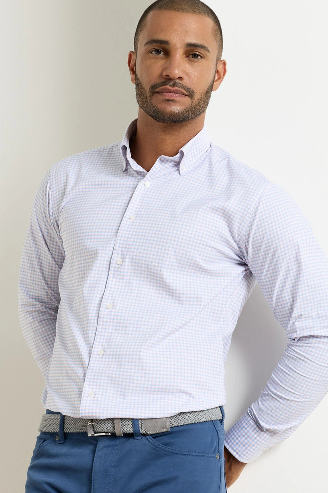 A man wearing a light blue and white micro-checkered long-sleeve button-down shirt, paired with blue trousers and a gray patterned belt, standing against a plain off-white background.