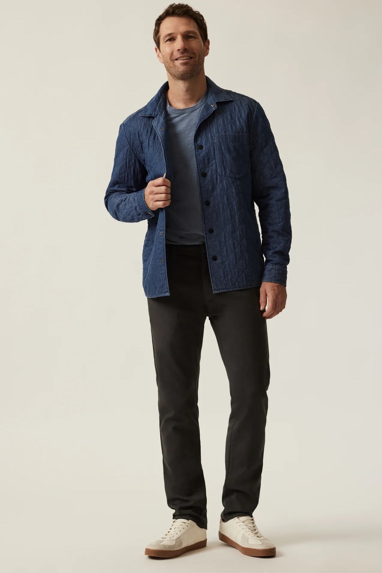 A man modeling a textured blue button-up overshirt over a dark gray t-shirt, black trousers, and off-white sneakers, photographed against a neutral light gray studio background.
