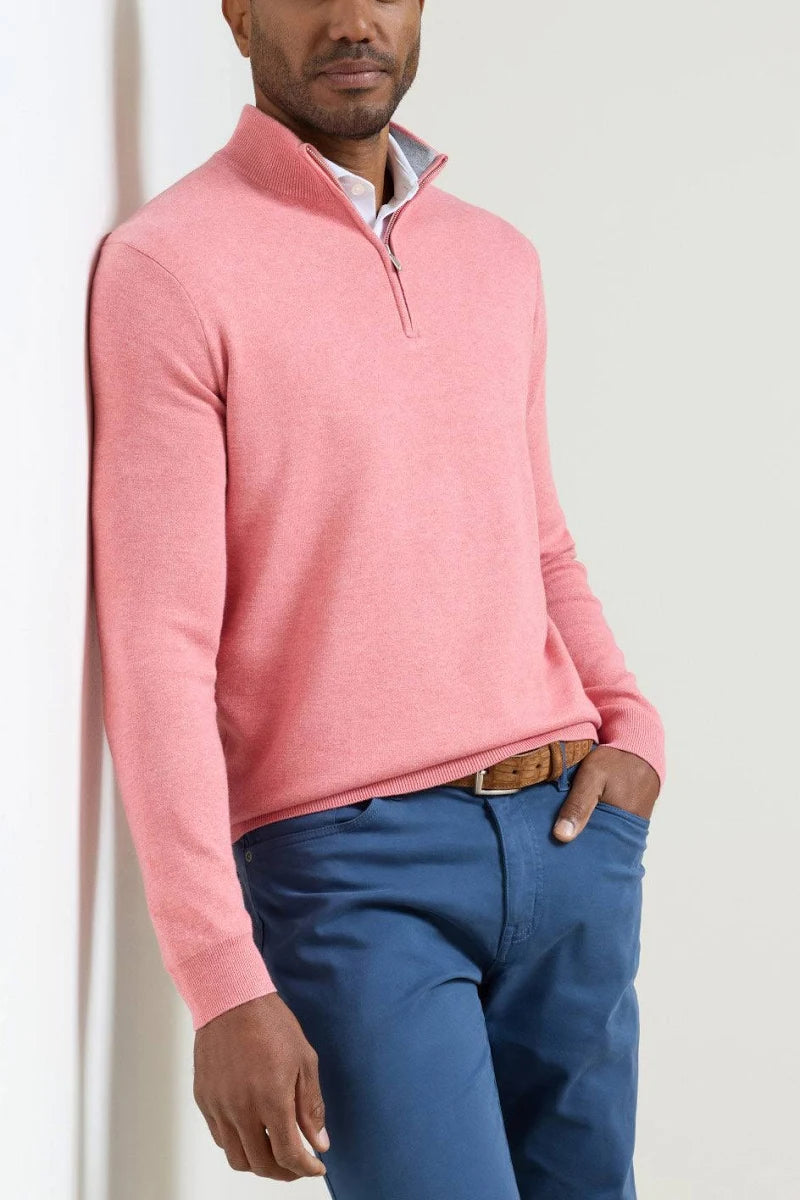 A man wearing a pink quarter-zip knit sweater over a light blue collared shirt and blue trousers, standing against a plain off-white wall.