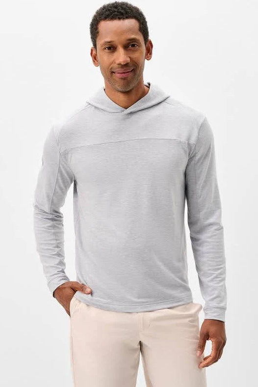 A man wearing a light gray long-sleeve hooded shirt and beige pants, standing against a plain white studio background.