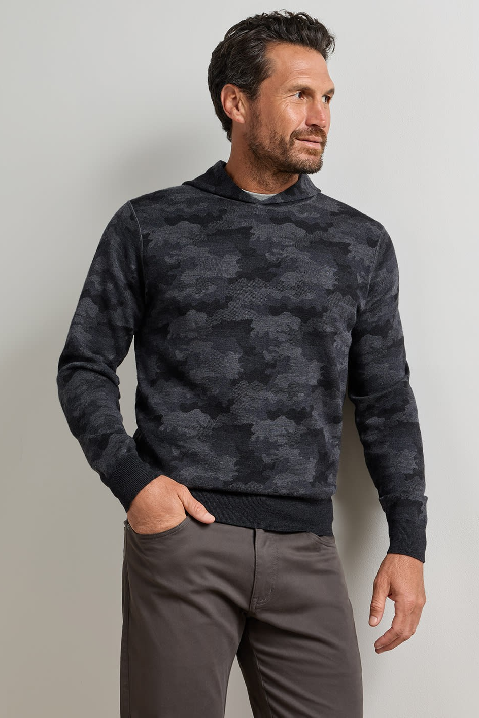 A man wearing a dark gray and black camouflage knit sweater with a ribbed hem, paired with gray trousers, standing against a light gray studio background.