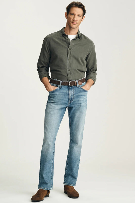 A man modeling a dark olive green long-sleeve button-down shirt, light blue jeans, a brown belt, and brown suede boots against a light gray studio background.