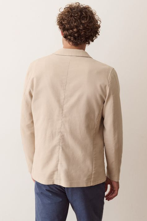 A man shown from the back wearing a light beige blazer and blue jeans against a plain off-white background.