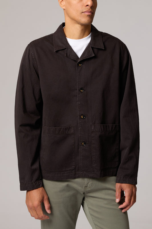 A man wearing a dark brown utility jacket over a white t-shirt and olive green pants, photographed against a neutral gray studio background.