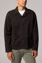 A man wearing a dark brown utility jacket over a white t-shirt and olive green pants, photographed against a neutral gray studio background.