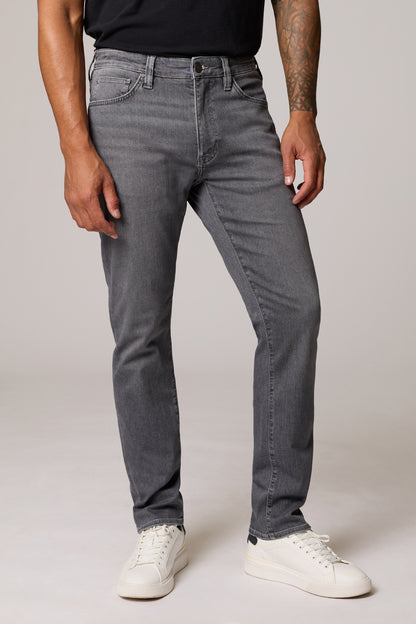 A man wearing slim-fit gray denim jeans, a black t-shirt, and white sneakers, standing against a light gray studio background.