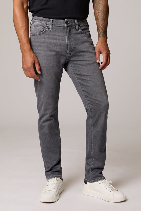 A man wearing slim-fit gray denim jeans, a black t-shirt, and white sneakers, standing against a light gray studio background.