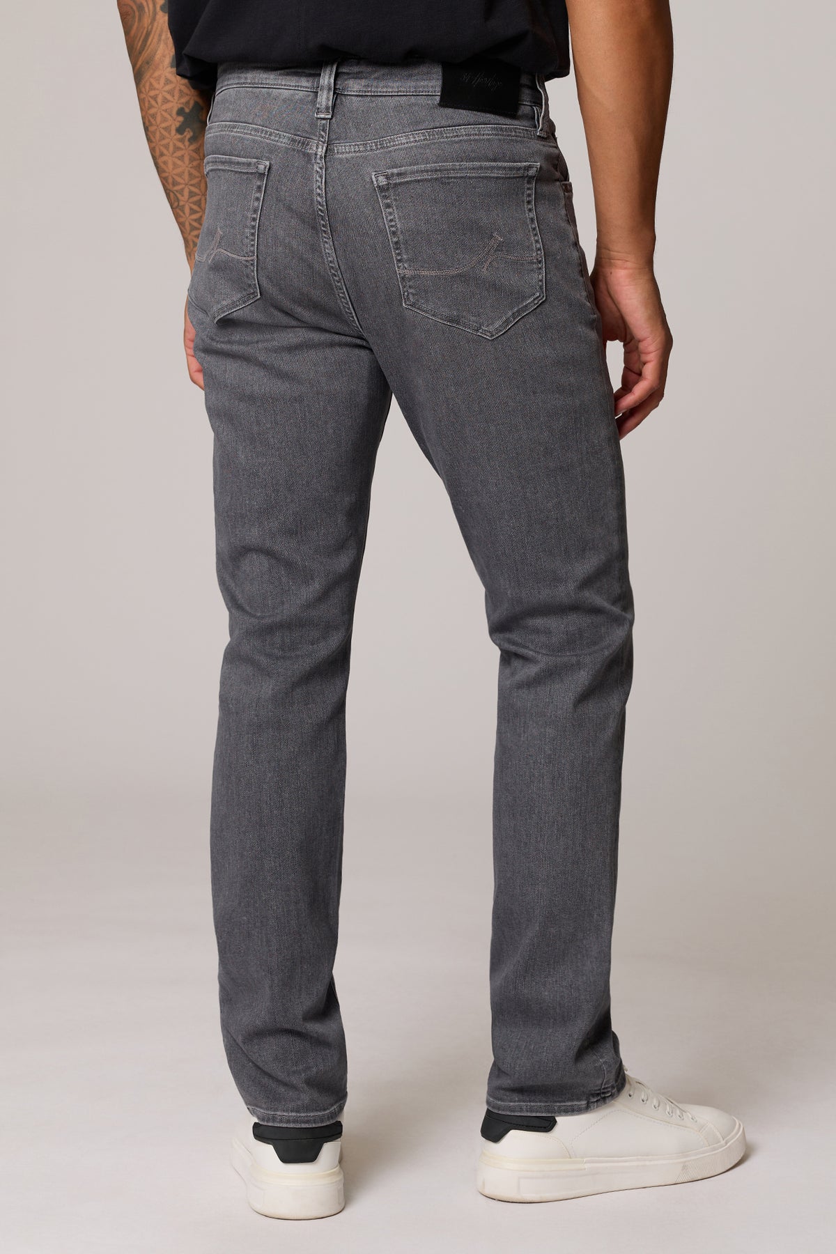 A person shown from the back wearing dark gray slim-fit jeans, a black t-shirt, and white sneakers against a light gray studio background.