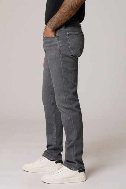 A side profile view of a person wearing dark gray slim-fit jeans, a black t-shirt, and white sneakers against a light gray studio background.
