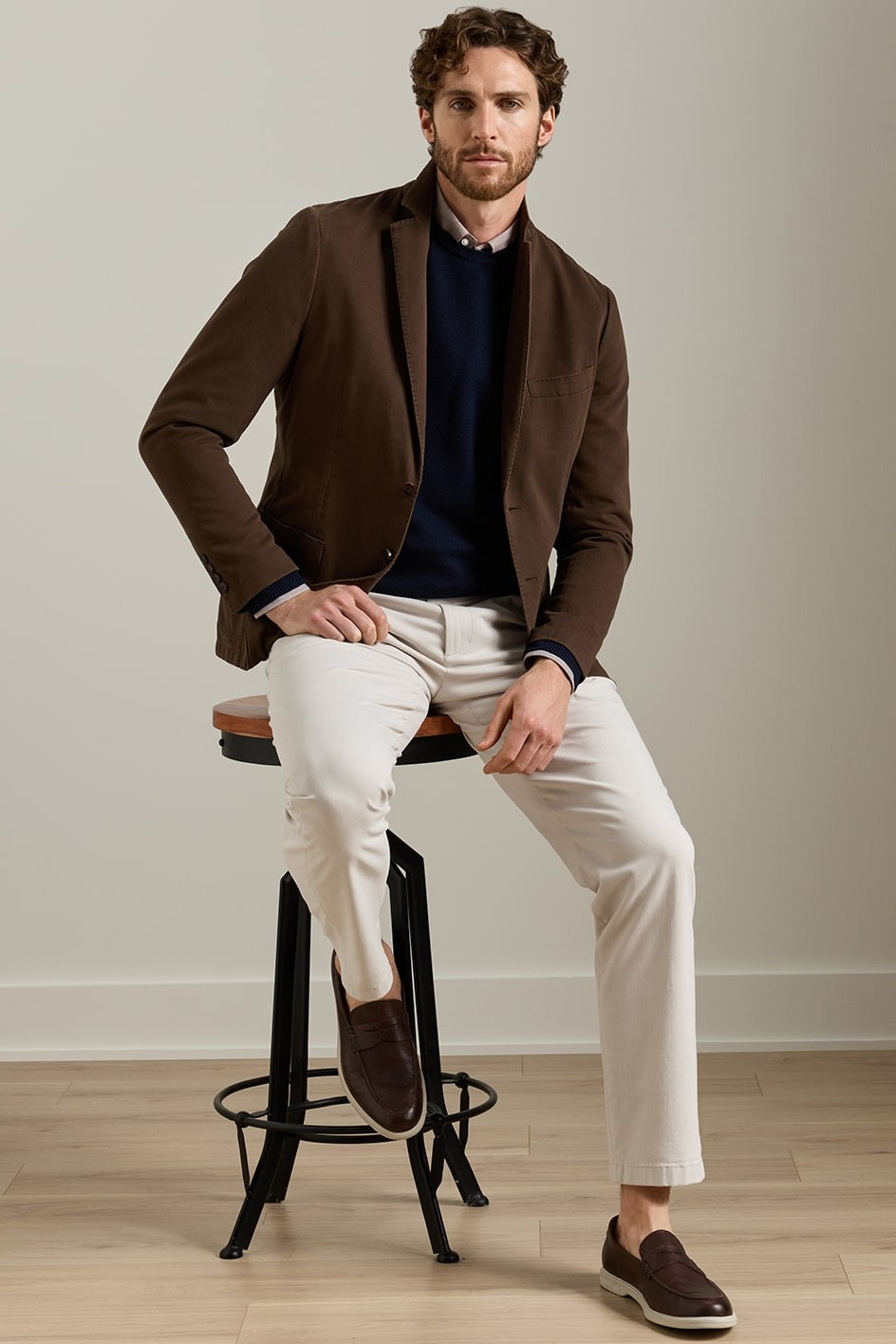 A man wearing a brown blazer, navy sweater, cream trousers, and brown loafers sits on a black metal stool in a studio setting with a light beige wall and wood floor.