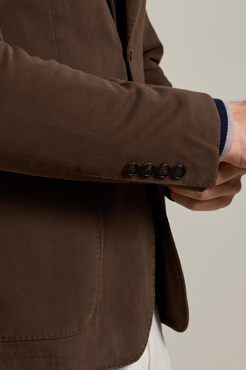 Close-up of a person wearing a dark brown textured blazer jacket, showing the sleeve with four buttons and a hand adjusting the cuff, set against a plain light gray background.