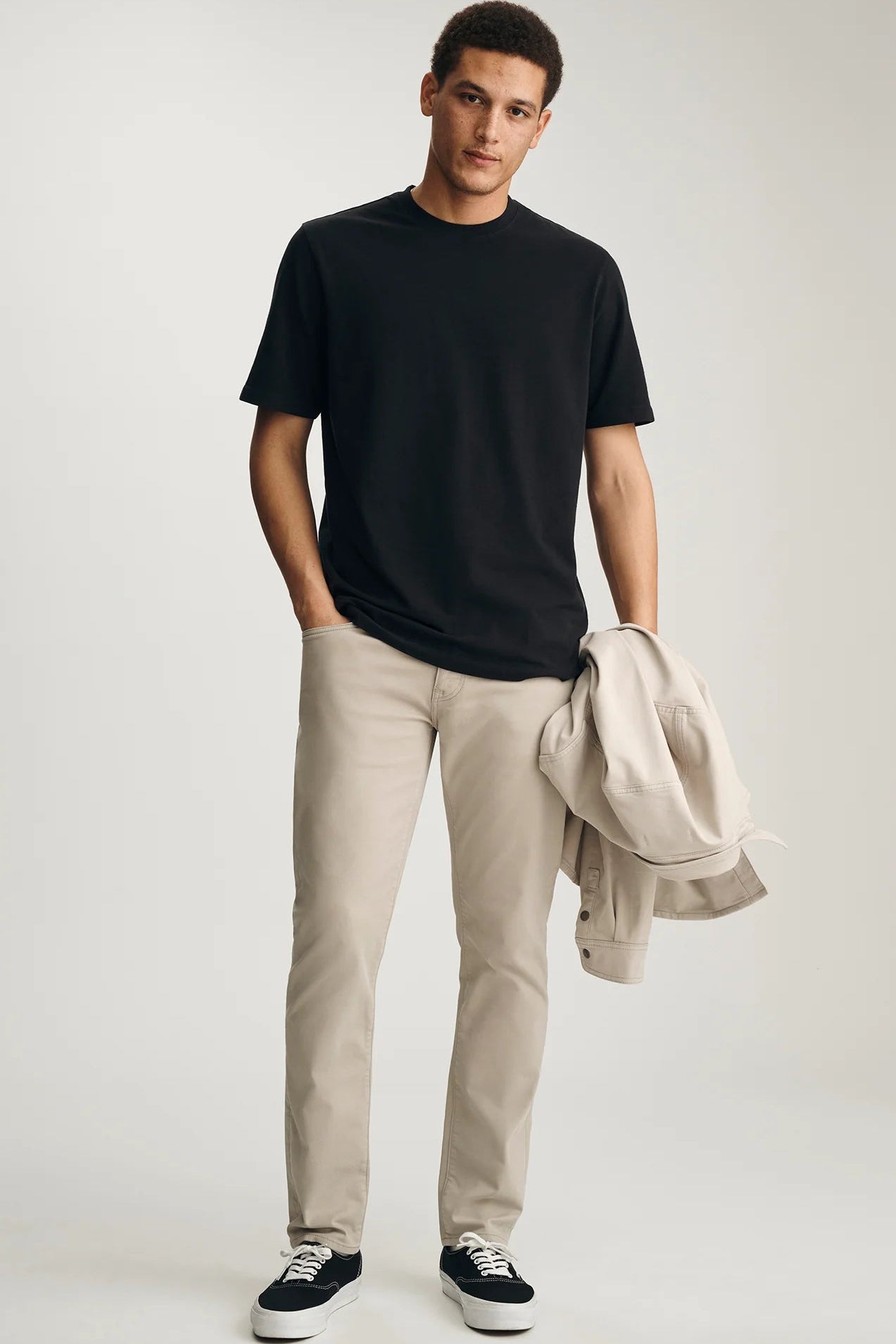 Man wearing a black t-shirt, beige pants, and black sneakers with a beige jacket over his shoulder on a white background.