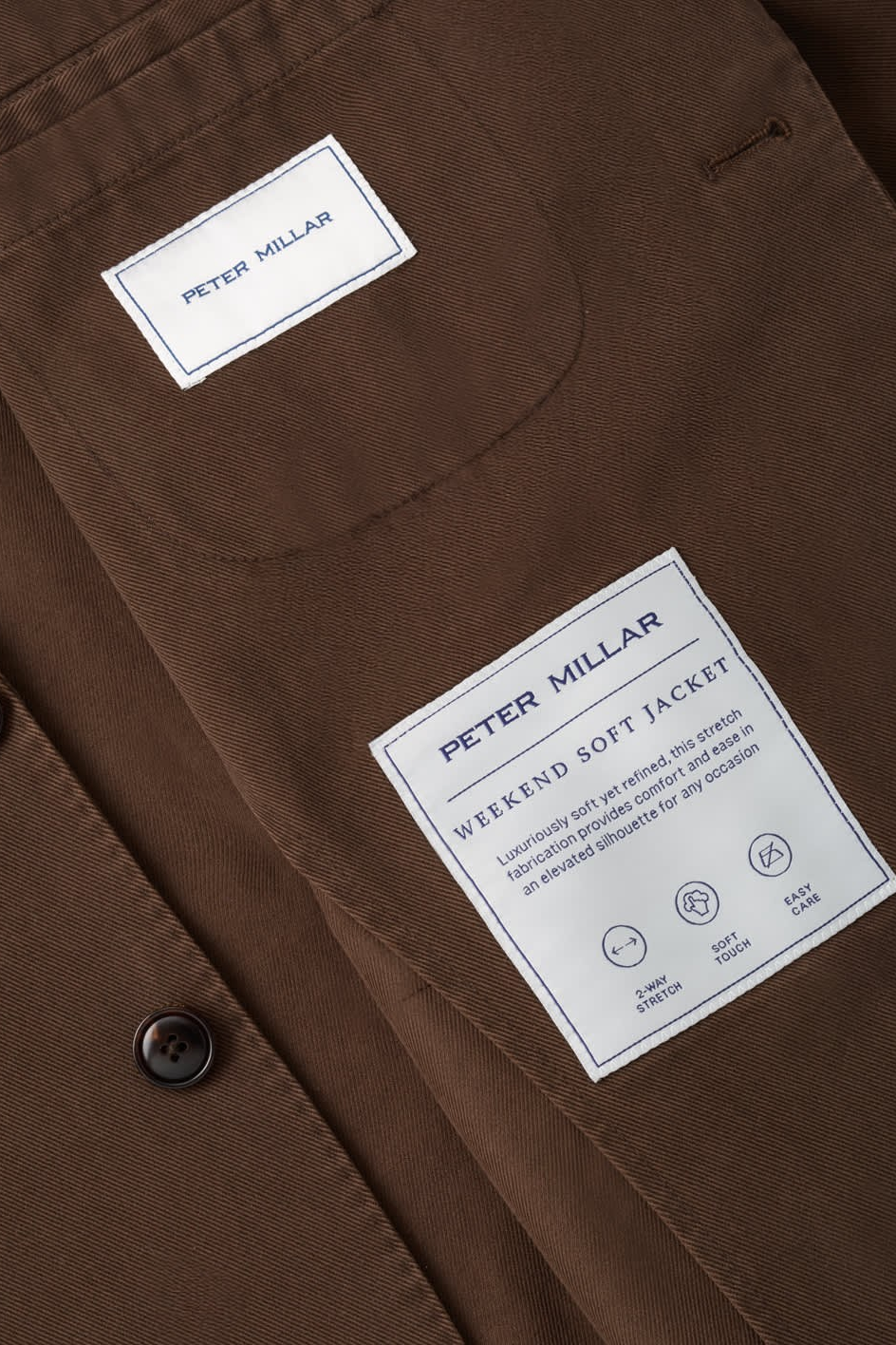 Close-up of a brown jacket fabric with a white Peter Millar brand patch and a buttoned pocket.