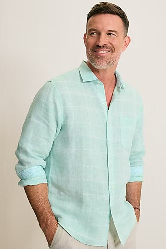 A man wearing a light mint green linen button-down shirt with rolled-up sleeves, paired with light beige trousers, standing against a plain off-white background.
